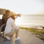 Un homme en chaise roulante et une femme regardent un coucher de soleil à la plage, l’homme désignant le soleil du doigt.