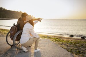 Un homme en chaise roulante et une femme regardent un coucher de soleil à la plage, l’homme désignant le soleil du doigt.